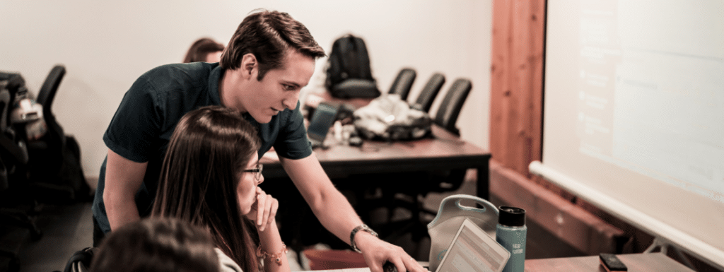 One white person standing next to another white person pointing at their laptop screen.