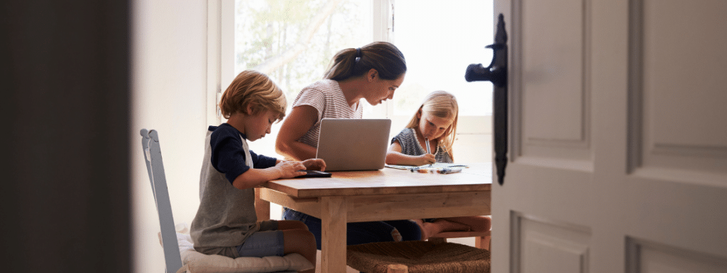 An adult sat at table with two children on either sides. The adult has a laptop open in front of them, one child is using a mobile device, while the other is holding a pen and whiting. 
