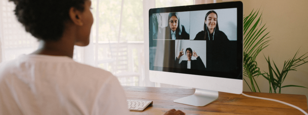 A person of color in front of a computer monitor engaged in a conference call. 