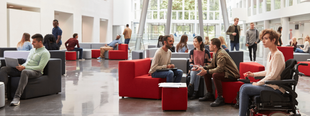 A lot of people in an open space, some seating on furniture in small groups talking, some standing or walking on the background. One person in a wheelchair passing by at the front. 
