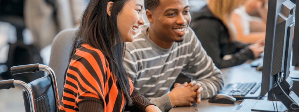 Two people seating at a desk and looking at same computer screen. 