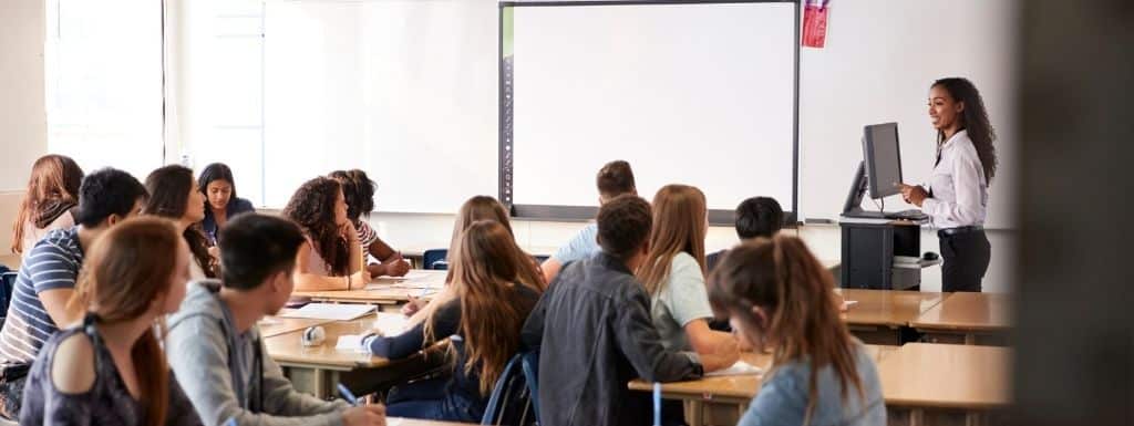 A classroom with students sat around tables in groups facing the teacher at the front. 