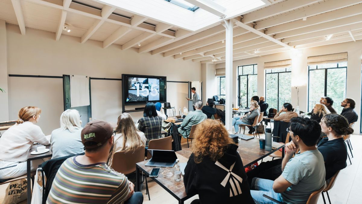 A group of people sat at desks looking at a screen with someone presenting at the front