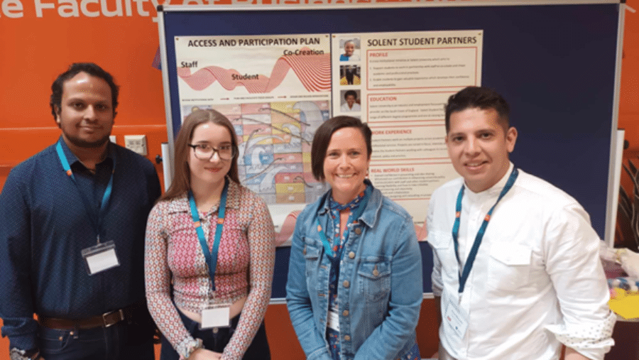 Four people stand smiling in front of a noticeboard display at a Southampton Solent University event. The display includes two posters: one titled "Access and Participation Plan" showing a co-creation diagram with interweaving staff and student pathways, and one titled "Solent Student Partners" outlining the programme's profile, education, work experience, and real-world skills. All four individuals are wearing lanyards with name badges.