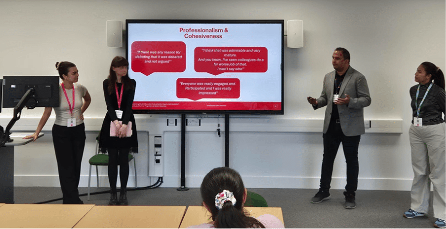 Four presenters stand at the front of a university classroom alongside a large screen displaying a slide titled "Professionalism & Cohesiveness." The slide features three red speech-bubble quotes from participants reflecting positively on group dynamics and engagement during a session. An audience member is visible in the foreground. The presentation appears to be from Southampton Solent University.
