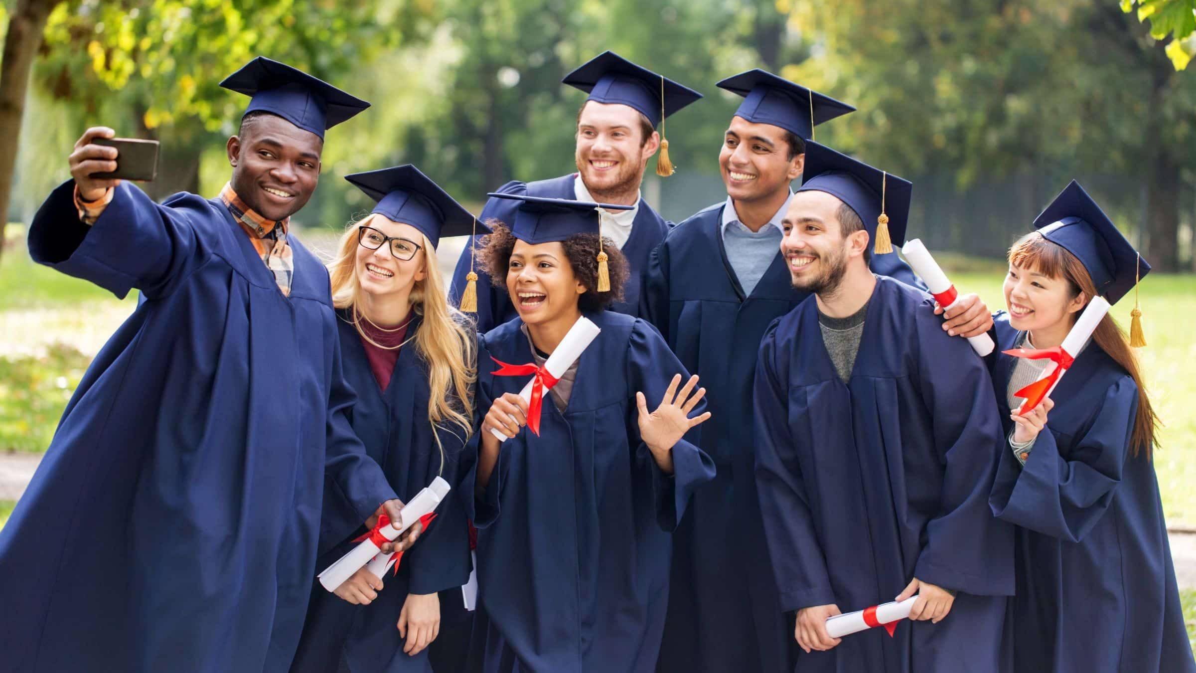 A diverse group of seven university graduates in navy cap and gown, smiling and taking a selfie outdoors, several holding rolled diplomas tied with red ribbon.