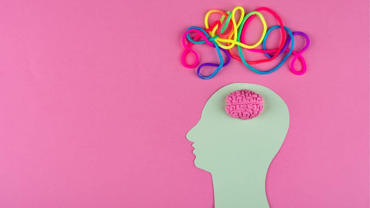 A pale green paper cutout of a human head in profile against a pink background, with a small pink brain model visible at the top of the head and a tangle of colourful ropes or cords emerging upward from it.