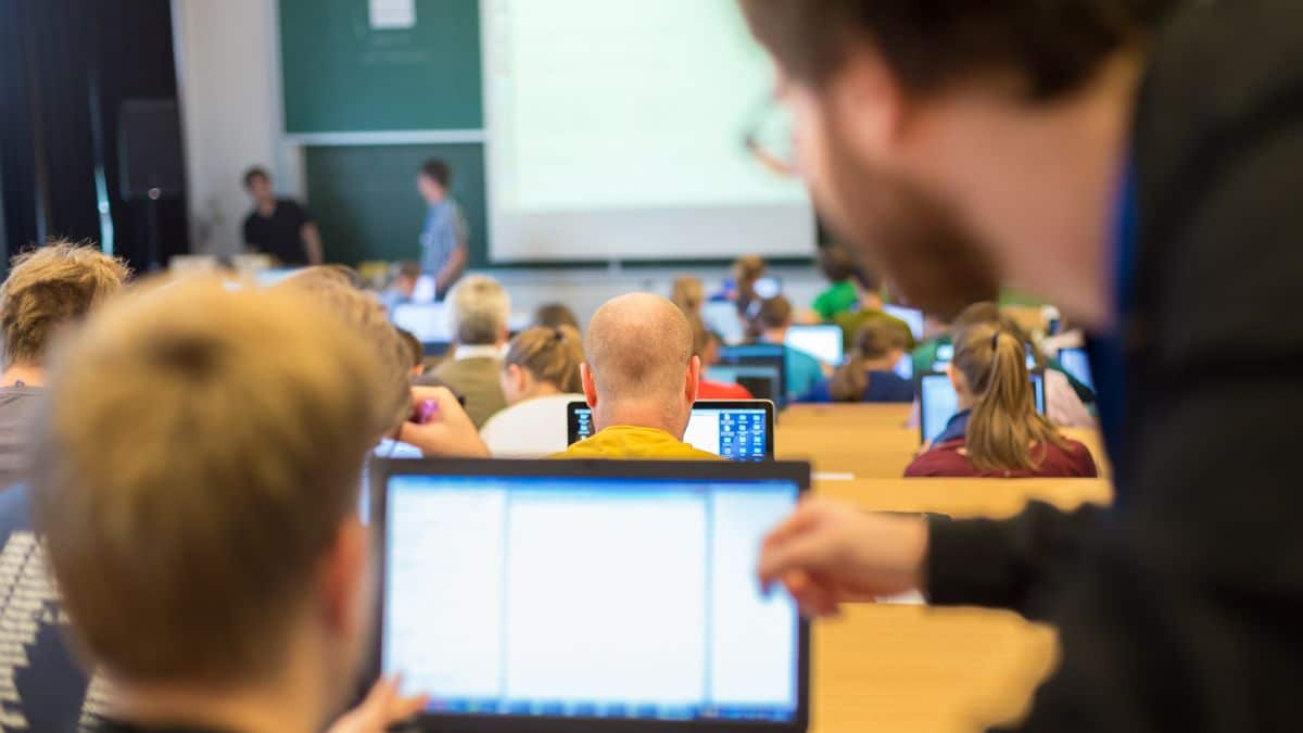 Lecture hall viewed from behind students working on laptops, with an instructor pointing toward a screen at the front of the room