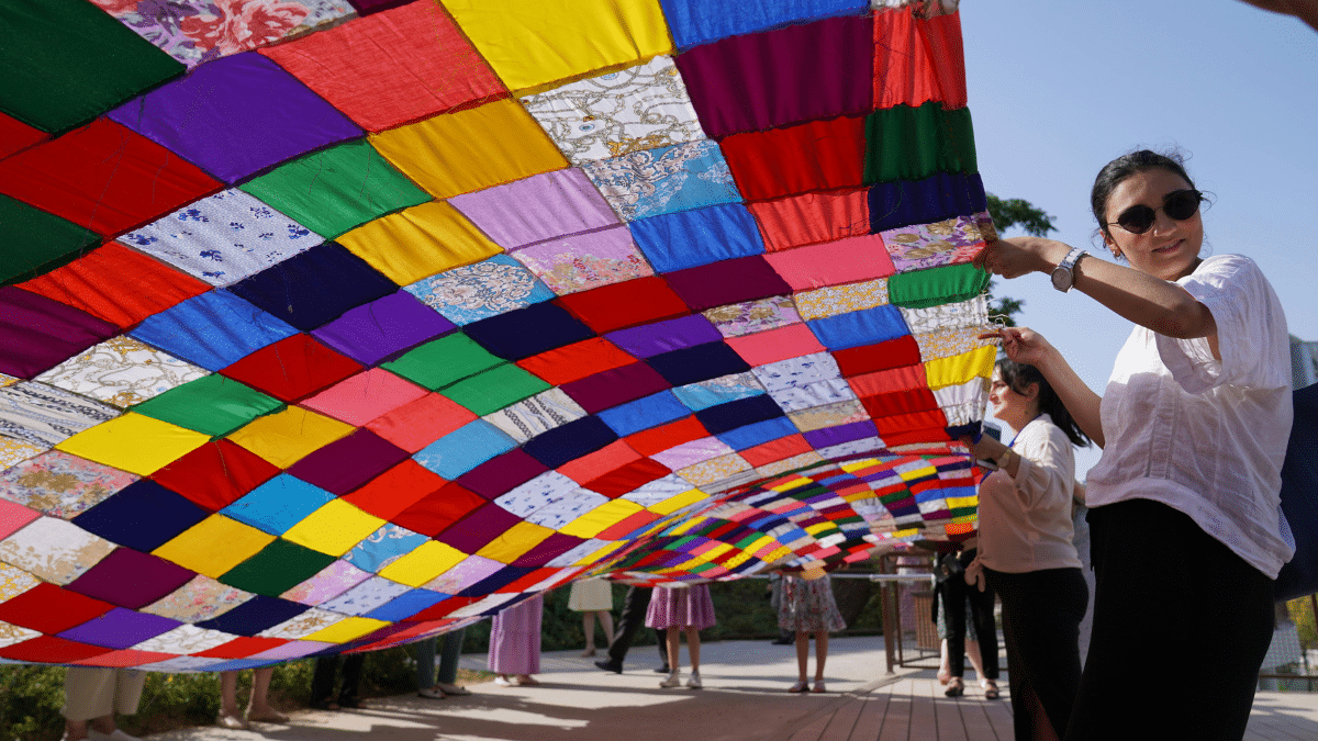 feature image: A colorful patchwork fabric canopy made of mixed-fabric squares stretches overhead outdoors, held up by a person in sunglasses and a white blouse in the foreground.