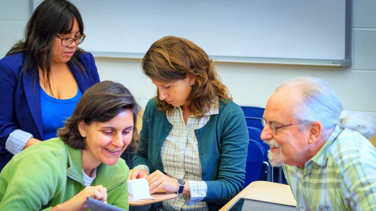 Feature image: Four colleagues gather around a desk in a classroom, smiling and looking at papers and a tablet together during a collaborative activity.