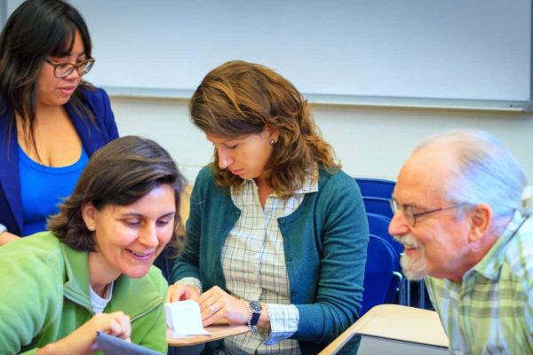 Feature image: Four colleagues gather around a desk in a classroom, smiling and looking at papers and a tablet together during a collaborative activity.