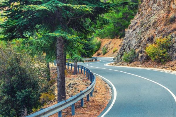 Feature image showing a winding mountain road with a metal guardrail curves through pine trees, with a rocky cliff face on the right side.