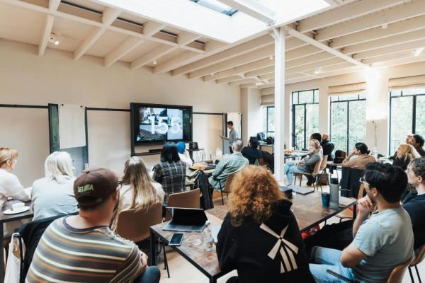 A group of people sat at desks looking at a screen with someone presenting at the front