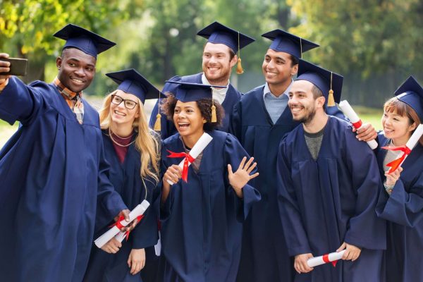 A diverse group of seven university graduates in navy cap and gown, smiling and taking a selfie outdoors, several holding rolled diplomas tied with red ribbon.