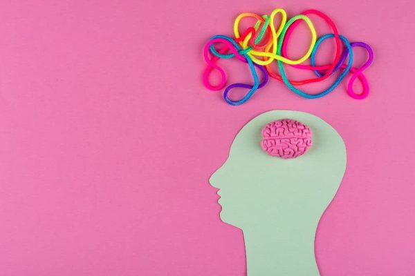 A pale green paper cutout of a human head in profile against a pink background, with a small pink brain model visible at the top of the head and a tangle of colourful ropes or cords emerging upward from it.